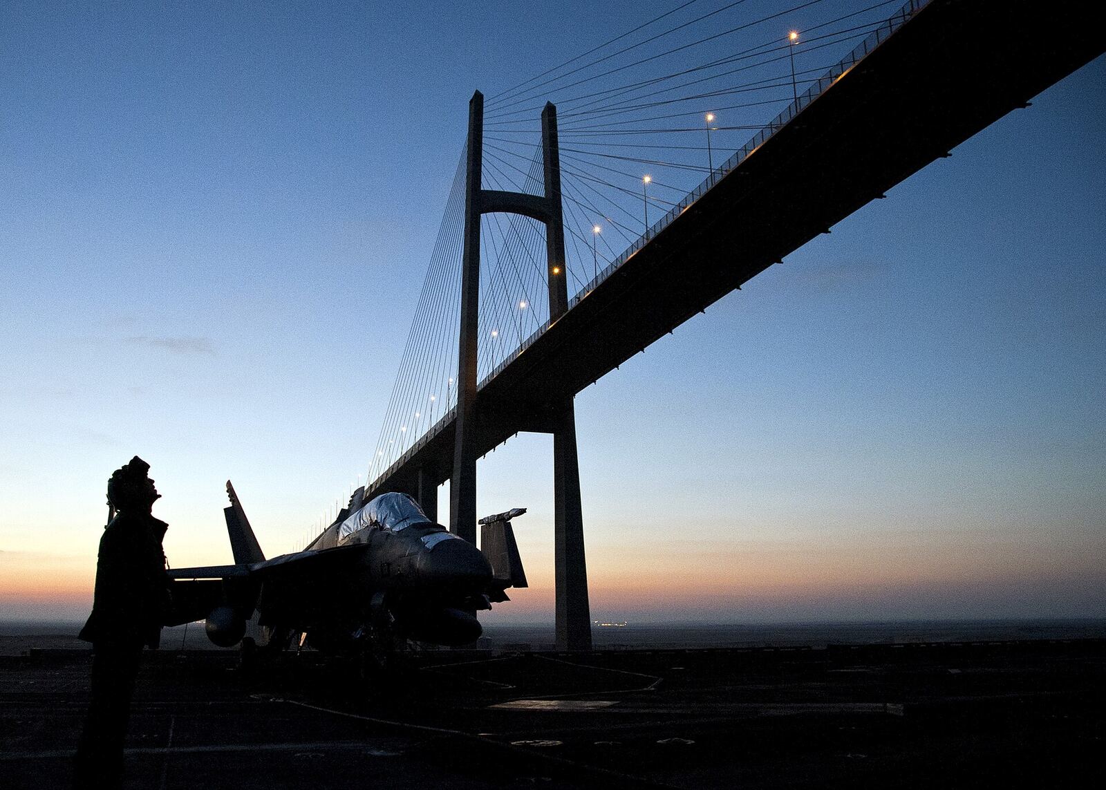 Fighter Jet | Love Hurtz A fighter jet rests on the deck of an aircraft carrier near a suspension bridge at dusk.
