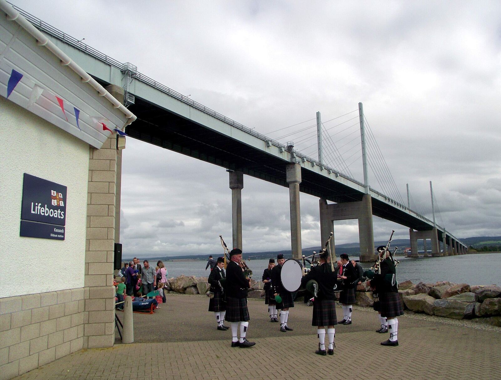 Forth Bridge | Love Hurtz A bagpipe band performs near the Forth Bridge and an RNLI lifeboat station, representing resilience through trauma.