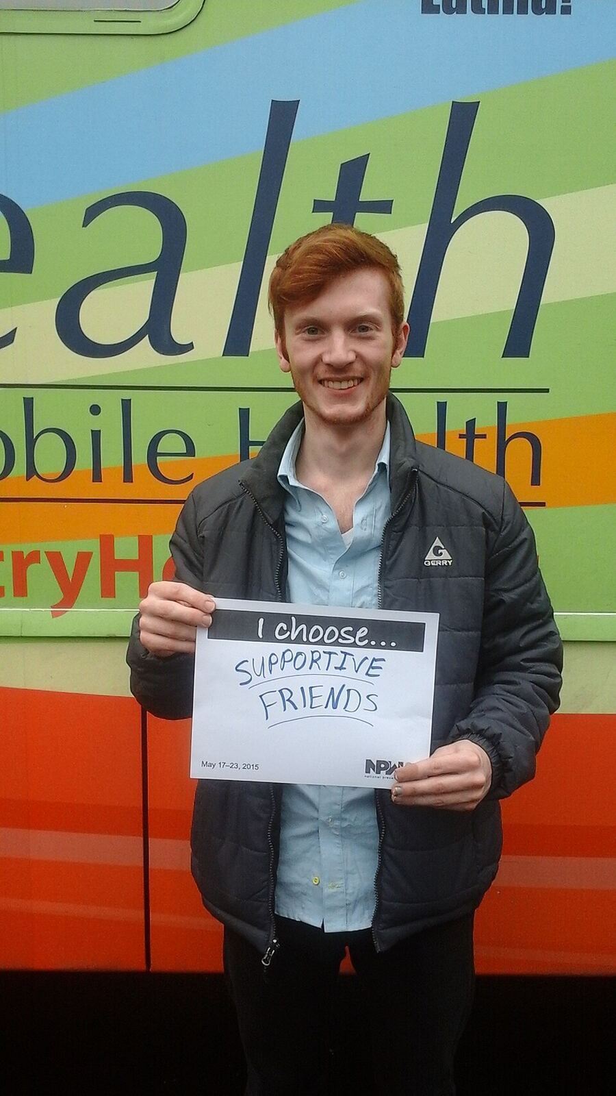 Man Promoting Mental Health | Love Hurtz A smiling red-haired man holds a sign promoting supportive friendships and mental health awareness.