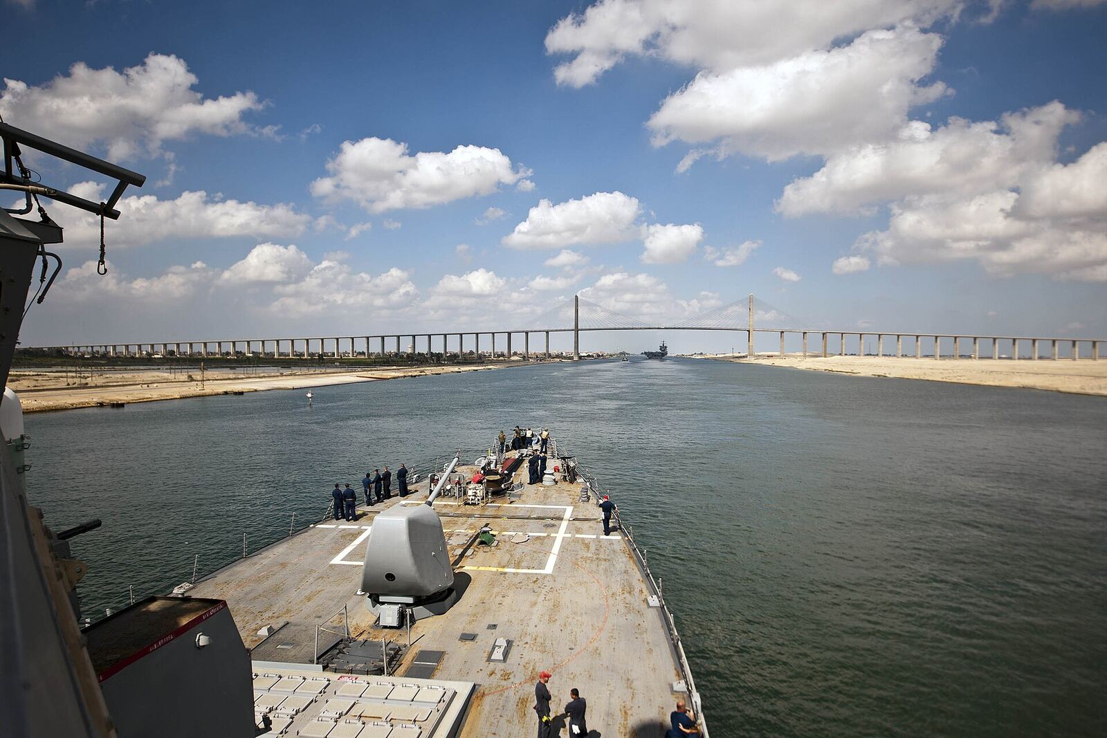 Suez Canal | Love Hurtz A naval vessel travels through the Suez Canal, a narrow waterway surrounded by desert landscapes and bridges.